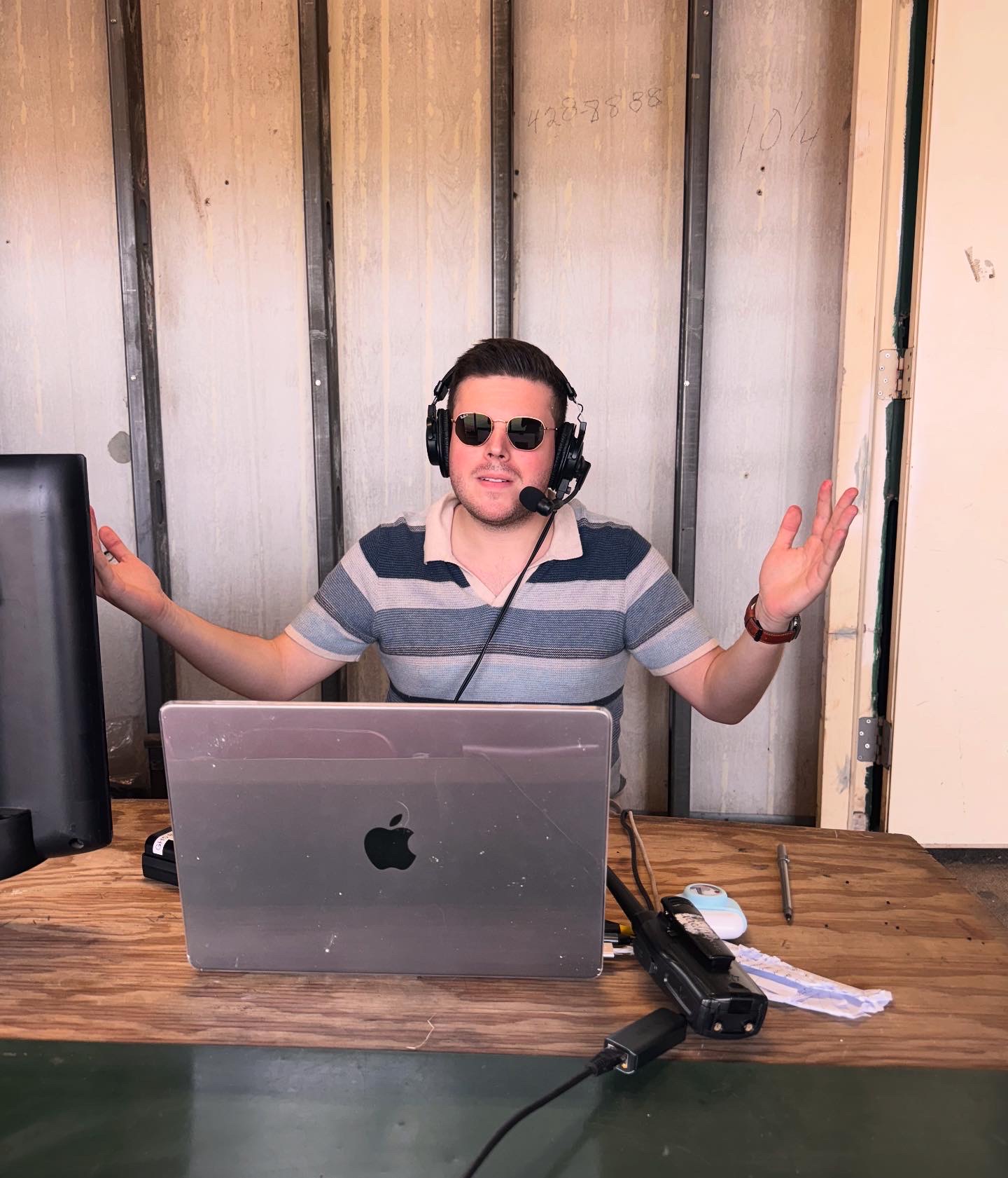 A man in a striped shirt and headphones stands behind a desk with a laptop, gesturing with his hands.